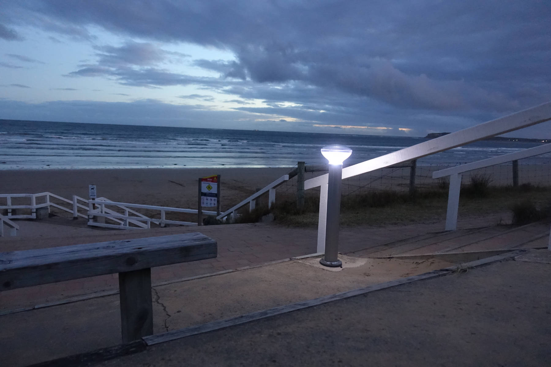 A solar bollard lighting the stairs and walkway at the entry to a coastal beach reserve at dusk
